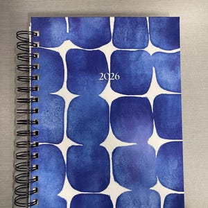 Peut inclure: Un agenda à spirales avec un motif bleu aquarelle composé de formes carrées arrondies. L'année "2026" est imprimée au centre. La couverture a un fond blanc avec des formes bleues.