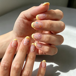 Può includere: Unghie di forma ovale con una manicure francese giallo chiaro su base trasparente. Ogni unghia è decorata con piccoli strass multicolori nelle tonalità blu, verde, arancione, viola e trasparente, sparsi sulla superficie dell'unghia.