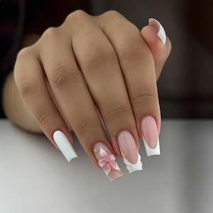 Può includere: Unghie artificiali lunghe di forma quadrata caratterizzate da un mix di smalto bianco solido, uno stile french manicure con punte bianche e un design floreale tridimensionale rosa e bianco accentuato da piccole perline metalliche dorate.