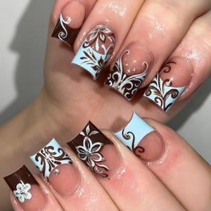 May include: Square-shaped artificial nails featuring a French manicure style with a light blue and dark brown colour palette. The design includes intricate white and brown floral patterns, swirling filigree details, and small embedded silver rhinestones.