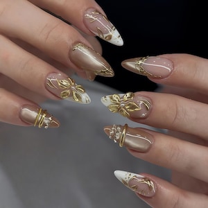 Könnte beinhalten: Nahaufnahme einer Hand mit langen, mandelförmigen Nägeln. Die Nägel weisen eine Kombination aus Gold- und neutralen Tönen auf, mit aufwendigen goldenen Blumen- und geometrischen Designs. Einige Nägel haben weiße Spitzen mit goldenen Akzenten.