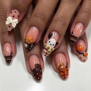 Op de afbeelding: Amandelvormige nagels met een nude basis en diverse herfstachtige 3D-kunst. De ontwerpen bevatten een witte strik, een pompoen, een klein figuurtje met een sjaal, koffiebonen, een stuk pompoentaart, oranje hartjes en zwarte stippen.