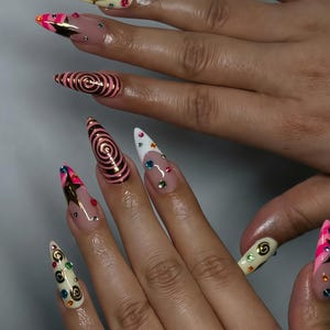 May include: Close-up of long, pointed fingernails with diverse designs. Some nails have a nude base with colourful gems, while others feature pink and gold accents. One nail has a spiral design, and another has a white tip with gems.