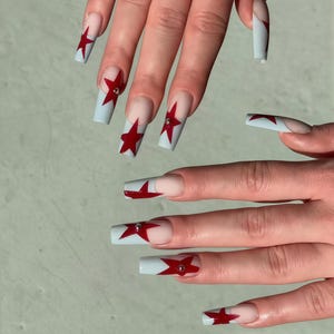 Könnte beinhalten: Künstliche Nägel mit hellblauem Grund und roten Sternen. Die Nägel sind lang und quadratisch, einige haben einen kleinen silbernen Edelstein. Das Design beinhaltet ein French-Tip-Design mit klarem Grund.