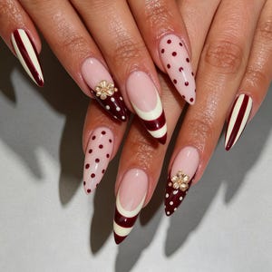 Puede incluir: Uñas en forma de almendra con una mezcla de diseños que incluyen rayas verticales en rojo oscuro y crema, lunares sobre una base rosa claro y puntas francesas con bandas crema y rojo oscuro. Dos uñas tienen pequeños adornos de flores doradas.