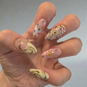 May include: A hand displaying five long, almond-shaped nails with intricate nail art. Designs include gold swirls, stars, and a spiral, along with colorful gems and a pink and white striped pattern. The nails have a glossy finish.