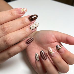 May include: Manicured fingernails featuring a variety of designs including brown leopard print, cream with gold polka dots, brown vertical stripes, and a white star shape with a small gold stud accent on a clear base, with one nail showing a brown tip.