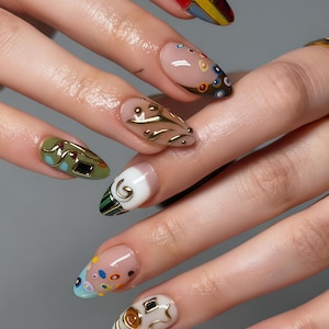 Könnte beinhalten: Nahaufnahme einer Hand mit mehreren langen, mandelförmigen Nägeln. Jeder Nagel hat ein einzigartiges Design, darunter geometrische Muster, goldene Verzierungen und bunte Punkte. Die Nägel sind in verschiedenen Farben lackiert, darunter Nude, Grün und Gold.