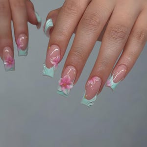 Puede incluir: Un conjunto de uñas acrílicas largas y cuadradas. Cada uña presenta una base transparente con un diseño floral rosa y blanco. Las uñas están acentuadas con puntas azul claro y pequeños adornos dorados.