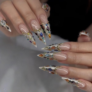 Op de afbeelding: Lange, puntige vingernagels met een nude basis en witte punten. Elke nagel heeft een goudkleurig metalen ontwerp met kleurrijke, juweelachtige accenten. Een bijpassende ring is ook zichtbaar.