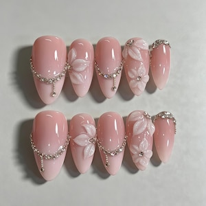 May include: A set of ten almond-shaped press-on nails in a soft, translucent pink gradient. The design features three-dimensional white floral accents, delicate silver-toned chains with small rhinestones, and clusters of clear crystals at the tips.