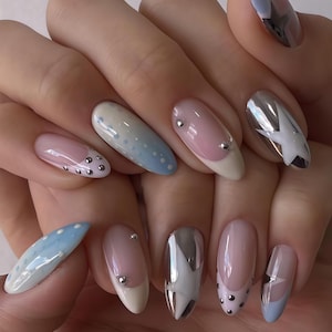 May include: Manicured nails with diverse designs. Some nails have a light pink base with a white tip and silver studs. Others feature a blue gradient with white dots, and some have a silver star design.