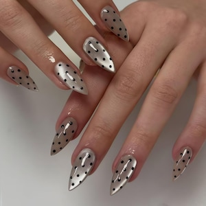 Op de afbeelding: Close-up van gemanicuurde nagels met een lichtbeige parelmoer afwerking. Elke nagel is versierd met kleine, gelijkmatig verdeelde zwarte stippen. De nagels zijn lang en puntig, wat een stijlvolle uitstraling creëert.