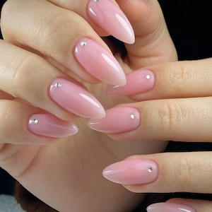 May include: Almond-shaped nails featuring a sheer, translucent light pink polish with a glossy finish. Each nail is accented with a single, small, clear rhinestone placed near the cuticle area, creating a clean and uniform aesthetic across all fingers.