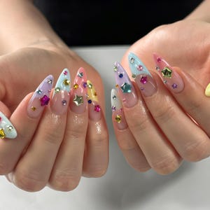 Puede incluir: Uñas en forma de almendra con estilo de manicura francesa y puntas de colores pastel en tonos azul, rosa, amarillo, lavanda y blanco. Las uñas están decoradas con varios pequeños dijes coloridos en forma de estrella y pedrería redonda.