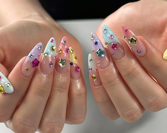Könnte beinhalten: Mandelformige Fingernägel mit French-Maniküre-Stil und pastellfarbenen Spitzen in Blau, Rosa, Gelb, Lavendel und Weiß. Die Nägel sind mit verschiedenen kleinen, bunten sternförmigen Anhängern und runden Strasssteinen verziert.