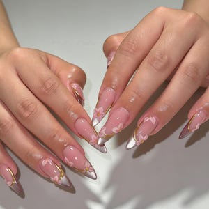 May include: Long almond-shaped nails featuring a sheer nude base with a metallic silver tip. Each nail is decorated with delicate pink floral accents and thin, curved gold metallic lines that frame the flowers and follow the nail shape.