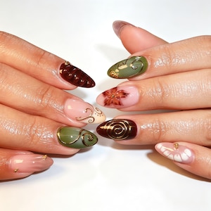 May include: Almond-shaped nails featuring a mix of olive green, deep brown, and soft pink polish. Designs include gold metallic swirls, a hand-painted brown flower, and small gold bead accents. Some nails have a solid colour finish while others feature intricate line work and textured patterns.