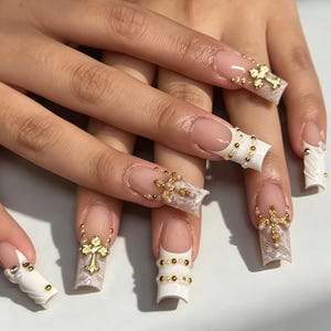 Puede incluir: Uñas postizas largas de forma cuadrada con base de manicura francesa y patrones de encaje blanco. Las uñas están decoradas con dijes de cruces doradas, pequeñas perlas blancas y diminutas cuentas metálicas doradas en las puntas.