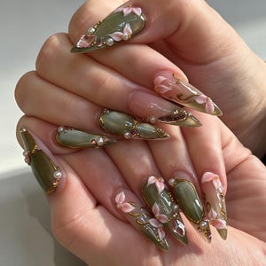 Può includere: Unghie a stiletto con una base verde oliva lucida, accenti metallici dorati, piccole perle bianche e decorazioni floreali rosa. Il design include sezioni trasparenti con volute dorate e cristalli rosa sfaccettati su diverse unghie.