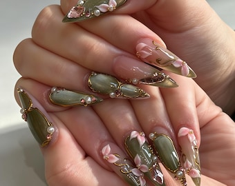Op de afbeelding: Stiletto-vormige nagels met een glanzende olijfgroene basis, gouden metalen accenten, kleine witte parels en roze bloemversieringen. Het ontwerp bevat transparante secties met gouden krullen en roze kristallen op verschillende nagels.