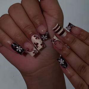 Op de afbeelding: Een manicure met lange, vierkante kunstnagels. De nagels tonen diverse ontwerpen, waaronder een French manicure met een donkerrode rand, bloempatronen met witte bloemen en afwisselende bordeauxrode en witte strepen. Sommige nagels hebben kleine, glinsterende versieringen.