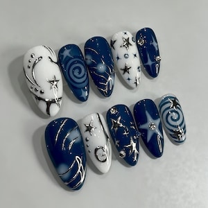 Peut inclure: Un ensemble de dix faux ongles en forme d'amande arborant un thème céleste avec des bases bleu marine profond et blanc. Les motifs incluent des étoiles métalliques argentées, des lignes tourbillonnantes et des strass incrustés sur certains ongles.