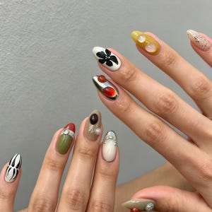 May include: Almond-shaped nails featuring a diverse manicure with varied colours including olive green, cream, mustard yellow, and white. The design incorporates 3D elements such as a black flower, metallic silver accents, red and brown cabochons, and small pearls.