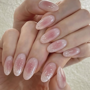 Pode incluir: Unhas de formato oval com uma base em degradê rosa suave que transita para um acabamento transparente e brilhante. As pontas das unhas são adornadas com partículas finas de glitter prateado cintilante que criam um efeito delicado.