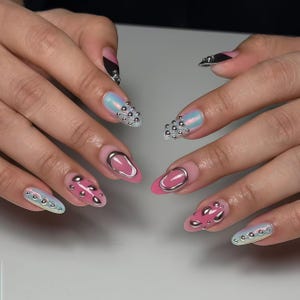 May include: Close-up of manicured nails with diverse designs. Some nails feature silver studs, others have pink and silver accents, and some have a metallic silver oval design. The nails are painted in shades of pink, iridescent blue, and black.