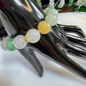 Könnte beinhalten: Ein Jade-Perlenarmband mit einer Mischung aus Farben, darunter Grün, Gelb und Weiß. Die runden Perlen sind mit goldfarbenen Verbindern aufgefädelt. Weitere Jade-Armbänder sind im Hintergrund zu sehen.