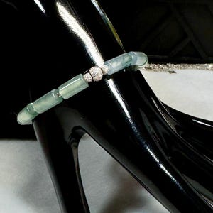 May include: A jade bracelet with a silver clasp and accents. The bracelet features rectangular jade beads in shades of green and white. The clasp is adorned with small, sparkling stones. The bracelet is displayed on a black surface.