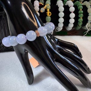 May include: A light purple jade bracelet with large, round beads. The bracelet is displayed on a black mannequin hand. A small, gold-colored spacer with red accents is between the beads. Other jade bracelets are visible in the background.
