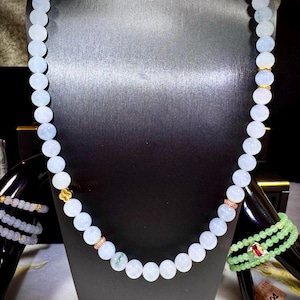 Può includere: Una collana di perline azzurre con dettagli dorati è esposta su un manichino nero. Un braccialetto abbinato è visibile a sinistra e un braccialetto verde con una gemma rossa è a destra. Le perline sono rotonde e di dimensioni uniformi.