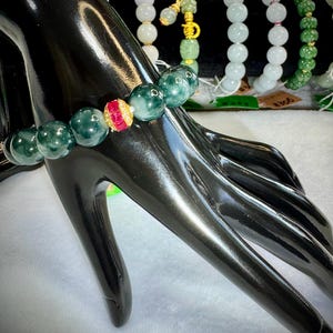 Könnte beinhalten: Ein dunkelgrünes Jade-Armband mit einer roten und goldenen Akzentperle. Das Armband wird auf einer schwarzen Mannequin-Hand präsentiert, mit anderen Jade-Armbändern im Hintergrund. Die Jadeperlen sind rund und poliert.