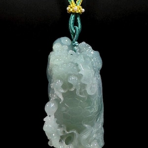 May include: A carved jade pendant in shades of green, suspended from a braided cord. The jade features intricate relief carvings, and the cord is green, yellow, and blue. The pendant is displayed against a black background.