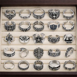 May include: A collection of silver-toned rings with various designs, including floral patterns, crosses, and star shapes. The rings are displayed in a wooden jewelry box with a beige interior, showcasing a variety of styles and sizes.
