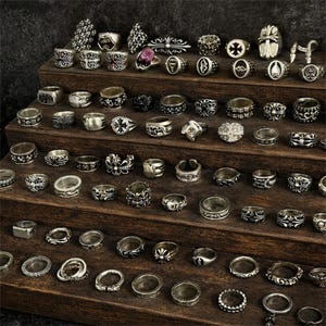 May include: A collection of silver rings displayed on a wooden tiered shelf. The rings feature various designs, including crosses, floral patterns, and geometric shapes. Some rings have gemstones. The rings are of different sizes and styles.