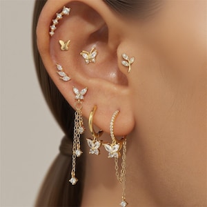Puede incluir: Una colección de pendientes y piercings dorados, que incluyen diseños de mariposas y hojas, adornados con piedras preciosas transparentes. Los pendientes incluyen aros y estilos de cadena colgantes, creando una estética delicada y elegante.