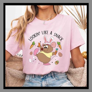 Peut inclure: T-shirt rose clair avec l'inscription "Lookin' Like a Snack" et une souris de dessin animé portant des oreilles de lapin et tenant des carottes. Des œufs de Pâques et des carottes entourent la souris.