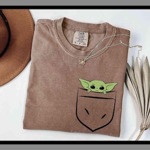 May include: A brown t-shirt featuring a green cartoon character peeking out of a pocket design. The shirt has a crew neck and short sleeves. A silver necklace with a small pendant is draped across the shirt.