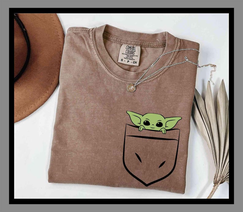 May include: A brown t-shirt featuring a green cartoon character peeking out of a pocket design. The shirt has a crew neck and short sleeves. A silver necklace with a small pendant is draped across the shirt.