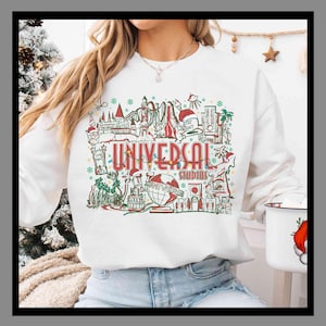 Może przedstawiać: Biała bluza z kolorową grafiką przedstawiającą atrakcje Universal Studios i czerwonym napisem "UNIVERSAL STUDIOS". Projekt zawiera różne zabytki i świąteczne akcenty, idealny dla fanów parków rozrywki.