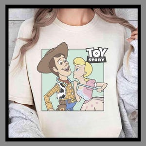 Puede incluir: Camiseta color crema con un gr&aacute;fico de dibujos animados de Woody y Bo Peep de Toy Story. Woody lleva un sombrero de vaquero y un chaleco, mientras que Bo Peep lleva un vestido rosa. Las palabras "TOY STORY" est&aacute;n impresas en negro sobre los personajes.