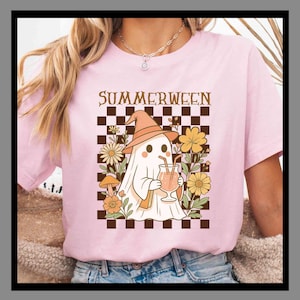 Può includere: T-shirt rosa chiaro con grafica "Summerween". Il design presenta un fantasma dei cartoni animati con un cappello da strega, che tiene in mano una bevanda e circondato da fiori. Lo sfondo è un motivo a scacchi neri e marroni.