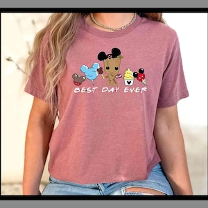 Könnte beinhalten: Ein mauvefarbenes T-Shirt mit einer Grafik von Disney-Figuren und Leckereien, darunter Mickey Mouse-Ohren, Zuckerwatte, Eis und Groot. Der Text "BEST DAY EVER" ist darunter aufgedruckt. Das T-Shirt wird mit blauen Jeansshorts getragen.
