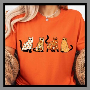 May include: Orange t-shirt featuring four cartoon cats in Halloween costumes. The cats are dressed as a ghost, a pumpkin, and other spooky characters. The shirt is a solid orange color.
