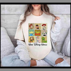 May include: A cream-coloured t-shirt displaying a grid of Toy Story character illustrations. The shirt features Woody, Mr. Potato Head, Jessie, Buzz Lightyear, Slinky Dog, Bo Peep, and Rex. The text "TOY STORY" and "Walt Disney World" are also printed on the shirt.