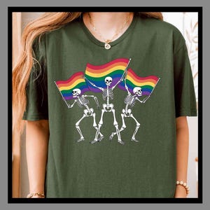 May include: Dark green t-shirt with a graphic of three skeletons holding rainbow flags. The skeletons are in a dancing pose. The flags have a rainbow design. The shirt is a solid colour.