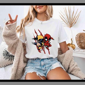 K&ouml;nnte beinhalten: Wei&szlig;es T-Shirt mit einer Cartoon-Grafik von Wolverine und Deadpool. Die Charaktere sind im Chibi-Stil, Wolverine in Gelb und Blau, Deadpool in Rot. Das Design beinhaltet rote Krallen, ein beliebtes Comic-Thema.